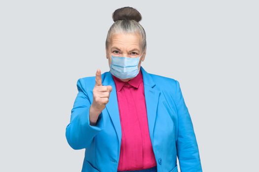 Angry aged woman with surgical medical mask warning you. Grandmother with light blue suit and pink shirt standing with collected bun gray hair. indoor studio shot, isolated on gray background