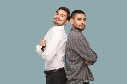 portrait of two handsome bearded successful friends or partners standing with crossed arms, looking at camera with proud face. profile, side view. indoor studio shot, isolated on light blue background