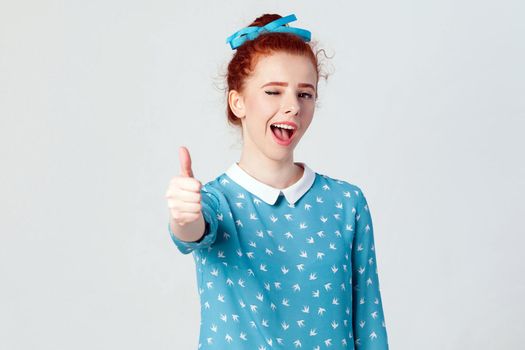 The young beautiful cheerful redhead girl showing thumb up and toothy smile and wink at camera. Isolated studio shot on gray background