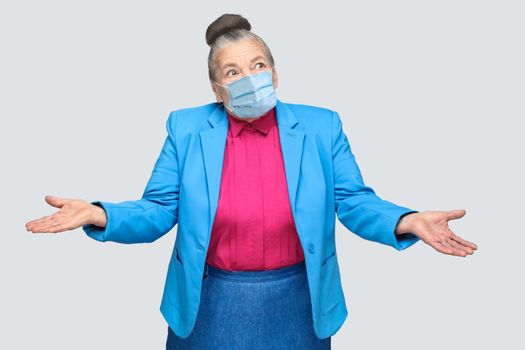 Puzzled or confused old woman with surgical medical mask, raised arms. grandmother with light blue suit and pink shirt standing with collected bun gray hair. indoor shot, isolated on gray background