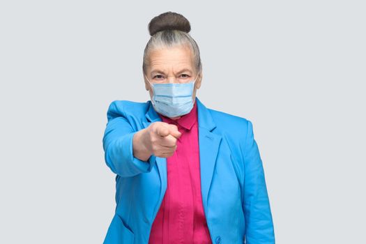 Hey you! Woman with surgical medical mask pointing at camera finger with serious face. Portrait of expressive grandmother standing with collected hair. indoor studio shot, isolated on gray background