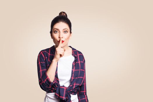 Portrait of beautiful young caucasian woman with black hair bun holding index finger at lips, asking to keep silence or not tell anyone her secret, raising brows, saying "Shh", "Hush", "Tsss"