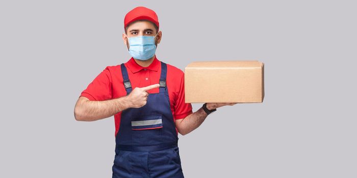 Delivery on quarantine. Young man with surgical medical mask in blue uniform and red t-shirt standing, holding and pointing finger to cardboard box on grey background. Indoor,studio shot, isolated,