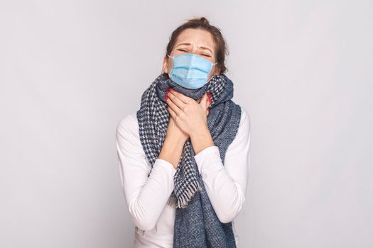 Sick young woman with surgical medical mask and blue scarf feeling bad and throat pain. indoor, studio shot, isolated on gray background