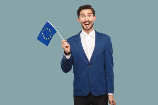 Time for Europe. Handsome amazed businessman in blue jacket and white shirt holding European union flag and looking at camera with wondered face. Indoor, studio shot isolated on light blue background.