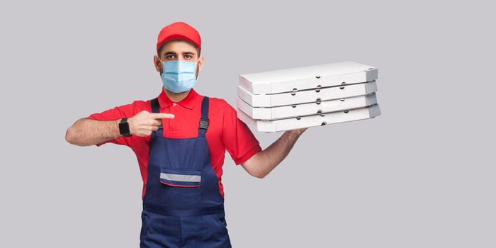 Delivery pizza on quarantine. Young man with surgical medical mask in blue uniform and red t-shirt standing, holding and pointing finger to stack of cardboard pizza boxes on grey background.