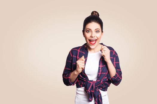 Portrait of pretty girl having winning and happy facial expression, exclaiming with joy, keeping hands in fists and mouth wide open, cheering, celebrating success or positive news, posing indoors.