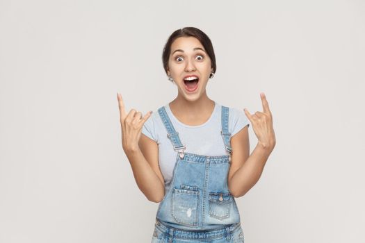 Rock n roll. Funny spanish woman in denim overalls, looking at camera with rock sing. Studio shot, on gray background.