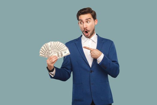 Handsome wondered businessman in blue jacket standing and holding many dollars in hand, pointing and looking at money with amazed surprised face. Indoor, studio shot isolated on light blue background.