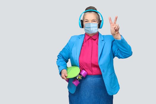 Aged woman with surgical medical mask in colorful casual style and blue headphones holding green skateboard listen music and showing peace sign, looking at camera. indoor, isolated on gray background