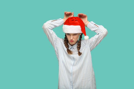 New year concept. funny beautiful girl in striped light blue shirt in red christmas cap standing with bunny gesture, winking, kissing and looking at camera. studio shot isolated on green background.