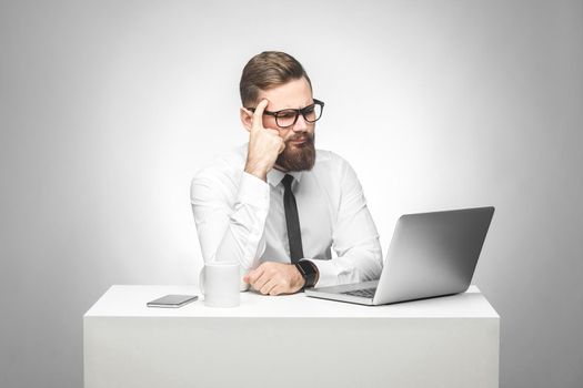 Thoughful bearded young boss in white shirt and black tie are sitting in office on desk and watching daily report on laptop, having new idea and planning own strategy, holding one hand on the head.