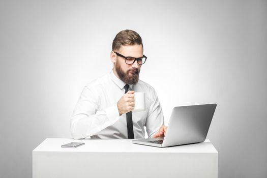 Coffee break! Portrait of handsome happy bearded young businessman in white shirt and black tie are sitting in office and have a break with cup of coffee and smile are looking to the laptop. Isolated