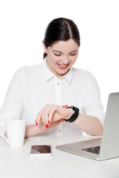 Portrait of happy attractive brunette businesswoman in white shirt sitting and looking at notification on her smartwatch, reading and toothy smile. indoor studio shot, isolated in white background.