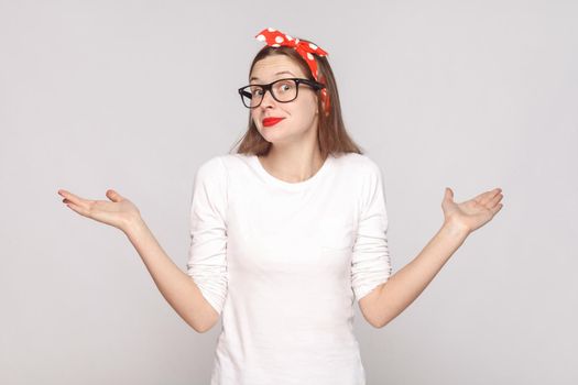 I dont know. i have no idea. portrait of beautiful emotional young woman in white t-shirt with freckles, black glasses, red lips and head band. indoor studio shot, isolated on light gray background.