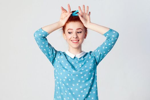 Stunning redhead girl with fabulous smile wearing blue dress holding hands on head pretending to be bunny. Isolated studio shot on gray background.