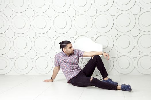 Young bearded fashion model in casual style is posing near white circle wall background. studio shot. sitting on floor and looking away.