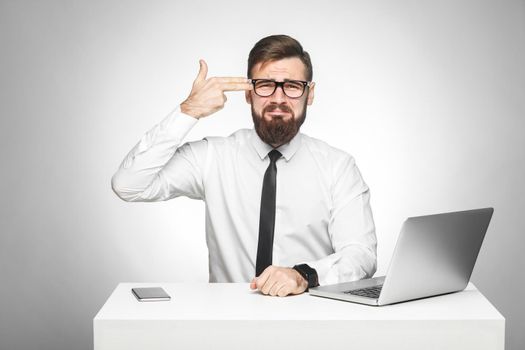 Unsatisfied stressed tired young manager in white shirt and black tie are sitting in office and trying to kill himself showing sign with finger pistol. Studio shot, isolated, gray background, indoor