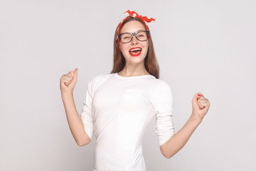 happiness and victory portrait of beautiful emotional young woman in white t-shirt with freckles, black glasses, red lips and head band. indoor studio shot, isolated on light gray background.