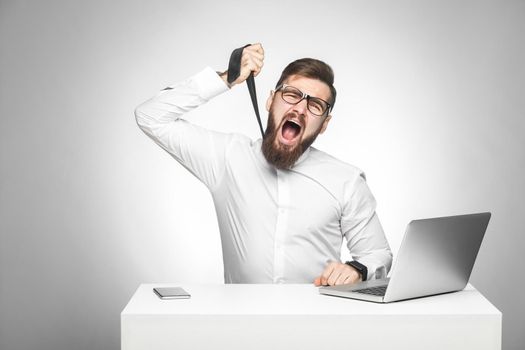 Portrait of unhappy stressed tired young manager in white shirt and black tie are sitting in office and trying to strangle himself with tie. Studio shot, isolated, gray background, indoor