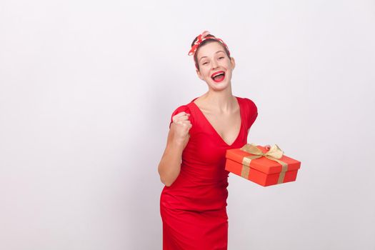 Yes! Win! Success beautiful woman holding gift box, toothy smile. Indoor, studio shot, isolated on gray background