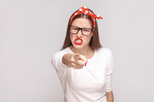 problem beause you are. anger pointing bossy portrait of beautiful emotional young woman in white t-shirt with freckles, glasses, red lips and head band. indoor, isolated on light gray background.