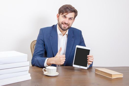 Technology, people and business concept - handsome man with beard and brown hair and blue suit and tablet pc computer and some books in the office, holding tablet and showing display screen with thumb. .Isolated on white background. .