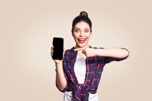 Young beautiful woman in casual style holding phone looking at camera and showing phone display with finger. studio shot on beige background.