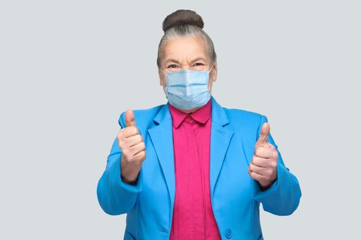 Aged woman with surgical medical mask showing like sign thumbs up. grandmother with light blue suit and pink shirt, collected bun gray hair. indoor s tudio shot, isolated on gray background