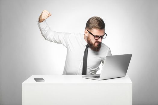 Portrait of aggressive unhappy businessman sitting in office and having bad mood are ready to punch a worker through a webcam with fist and angry face. indoor studio shot, isolated on grey background