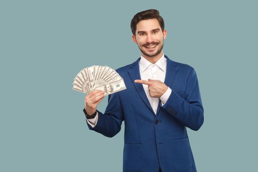 Handsome happy businessman in blue jacket standing and holding many dollars in hand and looking at camera with pointing finger and toothy smile . Indoor, studio shot isolated on light blue background