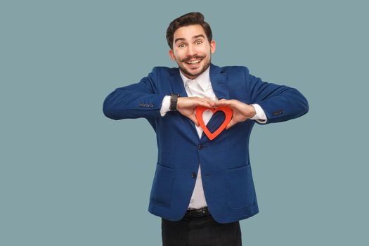 Handsome man in blue jacket and white shirt, standing and holding red heart shape and looking at camera with toothy smile. Indoor, studio shot isolated on light blue background