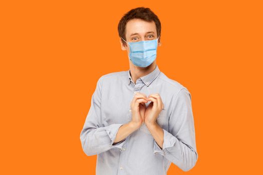 I love you. Portrait of joyful happy young worker man with surgical medical mask standing with heart love gesture and looking at camera with smile. indoor studio shot isolated on orange background.