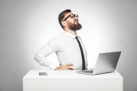 Side view of portrait of unhealphy upset young manager in white shirt and black tie are sitting in office and have strong backache or kidney pain. indoor, studio shot, isolated, gray background