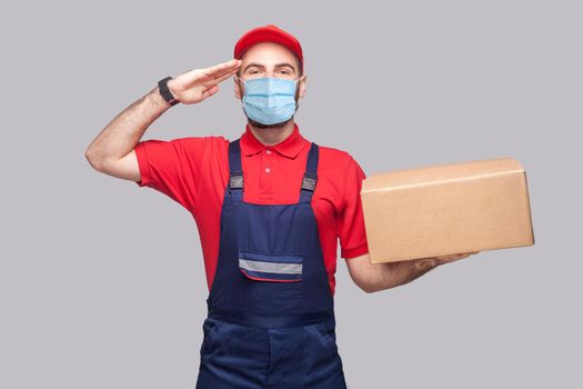 Delivery on quarantine. Give salute or yes sir! Young man with surgical medical mask in blue uniform and red t-shirt standing, holding cardboard box on grey background. Indoor, studio shot, isolated.