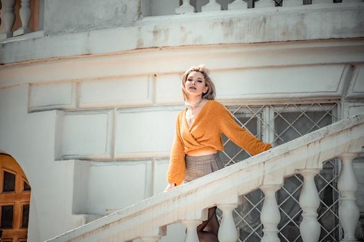 A middle-aged woman looks like a good blonde with curly beautiful hair and makeup on the background of the building. She is wearing a yellow sweater