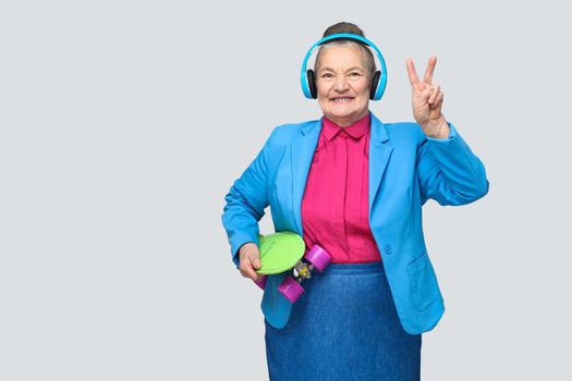 Trendy funny grandmother in colorful casual style with blue headphones holding green skateboard listen music and showing peace sign, looking at camera. Indoor, studio shot, isolated on gray background