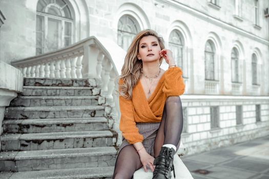 A middle-aged woman looks like a good blonde with curly beautiful hair and makeup on the background of the building. She is wearing a yellow sweater