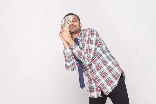 Dream come true! Satisfied rich handsome bearded businessman in colorful checkered shirt with blue tie standing and lean against to cheek fan of money. Indoor, studio shot, isolated on grey background