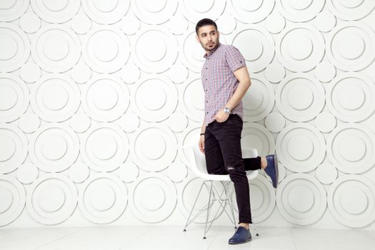 Young bearded fashion model in casual style is posing near white circle wall background. studio shot. posing on white chair and looking away.