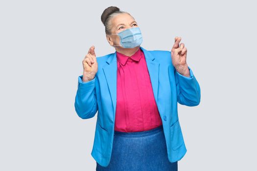 Hopeful with surgical medical mask woman crossed fingers and wish. Grandmother with light blue suit and pink shirt standing with collected bun hair. indoor studio shot, isolated on gray background