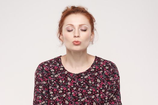 Portrait of ginger woman,closed eyes and send air kissing at camera. Studio shot, isolated on gray background