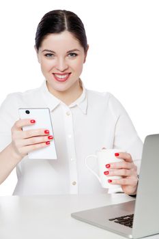 Portrait of happy successful attractive brunette businesswoman in white shirt sitting, holding cup of drink, smartphone and looking with toothy smile. indoor studio shot, isolated in white background.