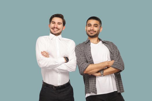 Portrait of two handsome satisfied bearded friends or partner standing with crossed hands and looking at camera with toothy smile. indoor studio shot, isolated on light blue background.