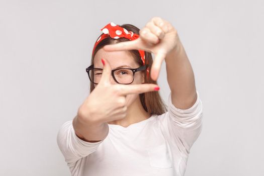 choosing composition or focusing portrait of beautiful emotional young woman in white t-shirt with freckles, glasses, red lips and head band. indoor studio shot, isolated on light gray background.