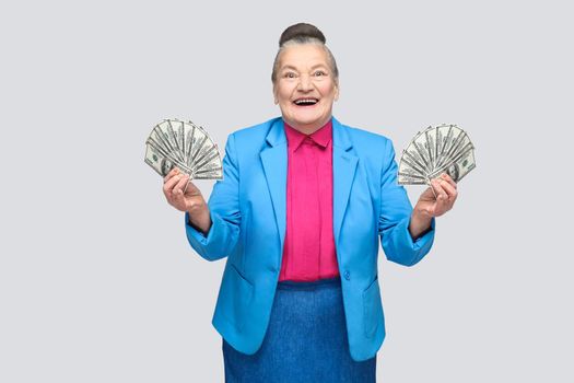 happy stylish aged woman holding many american dollars. Portrait of handsome expressive grandmother in light blue suit with collected gray hair bun hairstyle. Studio shot, isolated on gray background