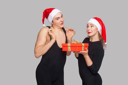 Woman present gift box man in santa hat. man with raised arms looking at camera with surprised face. New year concept. Studio shot, isolated on gray background