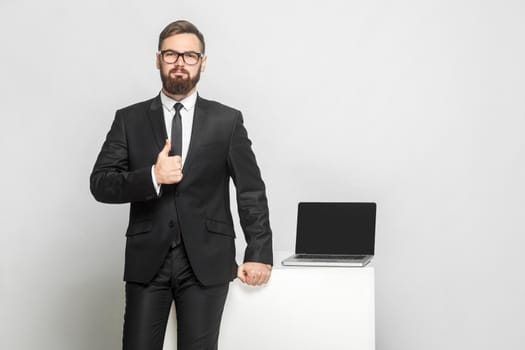 Portrait of handsome confident successful bearded young businessman in intelligence black suit are standing near his working place and showing thumbs up. Isolated, studio shot, indoor, gray background