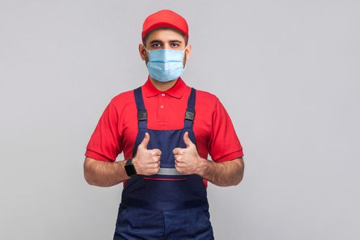 Work are done! Portrait of young man with surgical medical mask in blue overall, red t-shirt , cap, standing and showing thumps up and looking at camera. Grey background, indoor studio shot isolated.