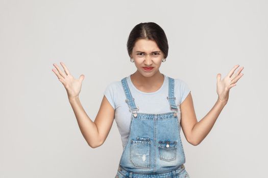 Anger spanish woman in despair and shock. Portrait of young adult angry woman in blue denimoveralls, looking panic in camera with mouth wide open, with hands up. Isolated on gray background.